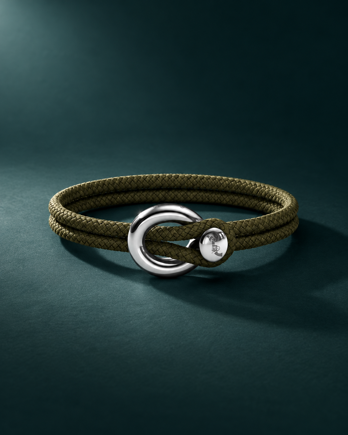 Imagine bracelet in olive cord and silver, photographed against a deep teal background. Editorial lighting.